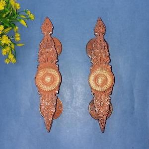 May include: Two ornate, bronze-colored door handles with intricate floral and scrollwork designs. Each handle features a central circular medallion with a sunburst pattern. The handles are mounted on circular backplates and are displayed against a blue background.