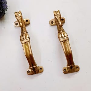 May include: A pair of antique brass door handles. Each handle has a horse head design at the top and a curved body. They are mounted on rectangular plates with screw holes. The brass has a warm, golden hue.