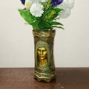 May include: An ornamental vase featuring a gold-coloured face design, filled with a bouquet of purple and white artificial flowers. The vase has a textured, antique look, and the flowers have green leaves.