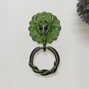 Peut inclure: Un heurtoir de porte en métal vert et noir représentant une tête de taureau. Le heurtoir a une poignée circulaire avec un motif de corde torsadée. La tête de taureau est entourée d'un motif floral décoratif.