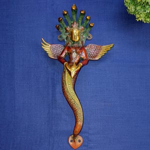 Puede incluir: Una detallada escultura de latón de una figura mítica con la parte superior del cuerpo humano, alas y una cola de serpiente. La figura tiene una corona, alas y está adornada con tonos rojos, dorados y verdes. La escultura está sobre un fondo azul.