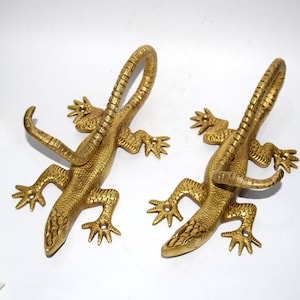 May include: Two gold-colored metal lizard hooks. The lizards are curled into a hook shape with their tails forming the hook. The lizards are detailed with scales and have a textured finish.