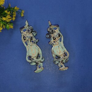 May include: Two matching metal door handles featuring an embossed figure of a dancing person. The handles have a dark bronze finish with light green patina accents in the recessed areas, showcasing intricate details on the clothing and jewelry.