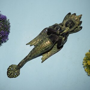 Op de afbeelding: Een groene en zwarte metalen deurklopper in de vorm van een mythisch wezen met vleugels. Het wezen heeft een mensachtig gezicht en houdt zijn handen bij elkaar in een gebedspositie.