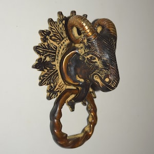 Puede incluir: Un llamador de puerta de latón con forma de cabeza de carnero con un anillo decorativo. La cabeza de carnero está detallada con cuernos, ojos y una cara texturizada. El anillo está retorcido y tiene un acabado dorado.