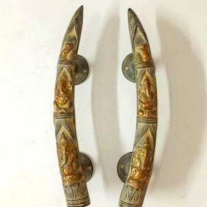 May include: Pair of antique brass door handles. Each handle is curved and features intricate carvings of a deity, with a gold finish. The handles are mounted on round, aged brass backplates, adding to their vintage appeal.
