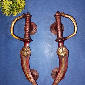 Puede incluir: Un par de tiradores de puerta ornamentados con un acabado dorado y rojo. Los tiradores tienen forma de espada curva con detalles intrincados.