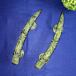 May include: Two ornate, curved door handles with a green and black finish. The handles feature intricate carvings and are mounted on circular, brass-colored plates. The background is a solid, deep blue fabric, with a small arrangement of green foliage in the upper right corner.