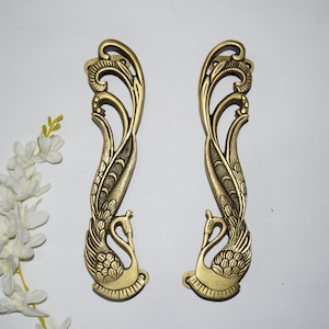 Puede incluir: Dos manijas de puerta doradas y ornamentadas. Cada manija presenta un diseño de pavo real estilizado, con el cuerpo del ave formando la base y sus plumas extendiéndose hacia arriba. Las manijas están sobre un fondo blanco.