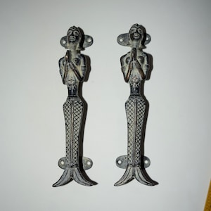 May include: A pair of antique-style door handles made of brass. The handles are shaped like women with their hands clasped in prayer. The handles are in a dark bronze finish.