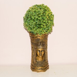 Könnte beinhalten: Eine goldene Vase mit einem ägyptischen Design, das ein Frauen Gesicht zeigt. Die Vase ist mit einer grünen Kunstpflanze gefüllt.