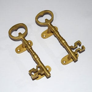 Puede incluir: Dos tiradores de puerta en forma de llave de color dorado con detalles ornamentados. Los tiradores están montados en placas rectangulares.