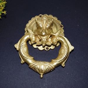 May include: A gold-toned door knocker with a detailed design featuring a mythical creature with a crown and a ring-shaped handle.