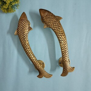 May include: A pair of gold-toned fish-shaped door handles. The handles are detailed with scales and have a curved shape.