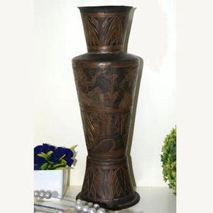 Könnte beinhalten: Eine hohe, antike Bronzevase mit aufwendigen Ätzmustern. Die Vase hat einen ausgestellten Sockel, einen breiten Mittelteil mit detaillierten Reliefschnitzereien und einen schmaleren, ausgestellten oberen Teil. Die dunkelbraune Farbe und die detaillierten Muster verleihen ihr eine Vintage-Ästhetik. Etwa 60 cm hoch.