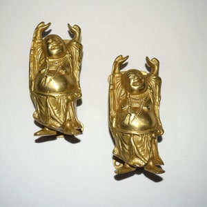 以下が含まれることがあります： 白い背景に立つ、笑う仏陀の金色の金属製置物2つ。それぞれが祝福のしぐさで腕を上げている。