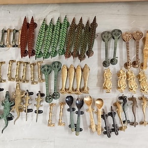 May include: A collection of decorative metal door handles in various designs and colors. The handles feature diverse shapes, including leaf, fish, and animal motifs, in shades of gold, green, brown, and black. These handles are likely for furniture or doors.