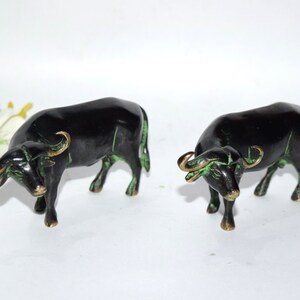 May include: Two small black metal buffalo figurines with green accents. The buffalo are standing on a white surface.