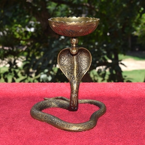 May include: A gold-coloured metal candle holder shaped like a cobra with intricate floral designs. The candle holder has a round bowl on top for holding a candle.