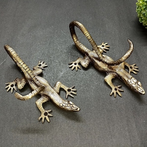 Puede incluir: Dos ganchos de pared de metal dorado en forma de lagarto. Cada lagarto tiene una cola enroscada que forma un gancho. Los lagartos están mirando en direcciones opuestas.