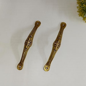 May include: A pair of antique brass cabinet pulls, each featuring a detailed face design. The elongated pulls have a central face motif and decorative lines. The brass has a warm, aged patina, indicating vintage craftsmanship. Each pull has mounting holes.