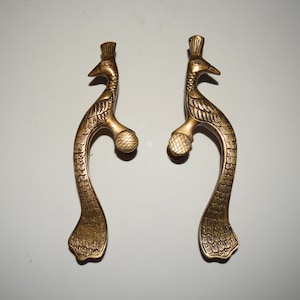 Könnte beinhalten: Zwei goldfarbene Türgriffe in Form eines Pfaus. Die Griffe sind mit aufwendigen Designs verziert und haben oben einen abgerundeten Knopf.