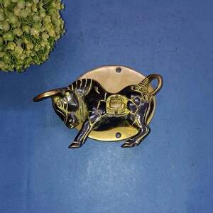 May include: A brass bull-shaped door knocker with a black body and gold accents. The bull is mounted on a gold-colored circular plate with three holes. The knocker is set against a blue background, with a cluster of green flowers in the upper left corner.