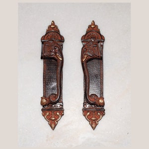 May include: A pair of ornate door handles featuring elephant head designs. The handles are made of dark brown metal with gold accents. The handles are mounted on a white wall.