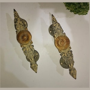 May include: Two ornate, decorative door handles with a gold and silver finish. The handles are shaped like a stylized leaf with a circular, gold center.