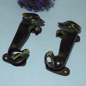 Op de afbeelding: Twee zwarte dackel deurgrepen met groene en gouden accenten. De grepen zijn van metaal en hebben een vintage uitstraling.