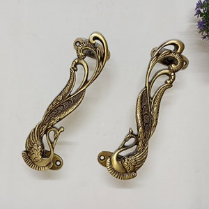 Puede incluir: Dos tiradores de puerta de latón adornados con forma de pavo real con detalles intrincados. Los tiradores son de color dorado y tienen un diseño decorativo.