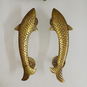 May include: Two brass door handles shaped like fish. The handles are gold-colored with detailed scale patterns and fin designs. The fish-shaped handles are mounted on circular plates, adding a decorative touch to any door.