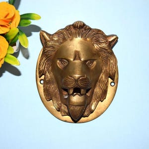 May include: A brass lion head door knocker with a detailed mane and facial features. The knocker is mounted on a circular plate with two screw holes. The lion's mouth is slightly open, revealing teeth. The background is a light blue.