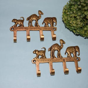 May include: Two brass-colored wall hooks, each with three hooks. Each hook features a decorative design of a monkey, camel, and elephant. The hooks are mounted on a light blue wall.
