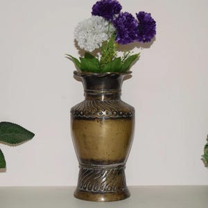 Könnte beinhalten: Eine bronzefarbene Vase mit dekorativem Design, die einen Strauß künstlicher Blumen enthält. Die Blumen umfassen weiße und violette Blüten mit grünen Blättern. Die Vase hat einen breiten Boden und eine ausgestellte Oberseite.