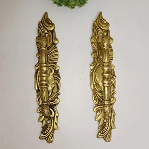May include: Two ornate, gold-colored door handles with a detailed, floral design. Each handle features a central, cylindrical grip and is flanked by elaborate, symmetrical embellishments. The handles are likely made of metal and are set against a white background.