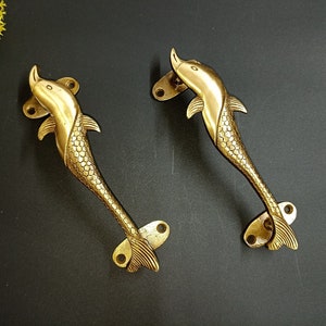 Könnte beinhalten: Zwei goldfarbene Delfin-Türgriffe mit einer strukturierten, schuppigen Oberfläche. Die Griffe sind mit einem gebogenen, delphinartigen Körper und einer Schwanzflosse gestaltet.