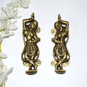 Puede incluir: Un par de tiradores de puerta de metal dorado con forma de figuras danzantes. Las figuras son de estilo indio tradicional, con ropas fluidas y detalles intrincados.