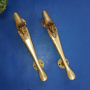 May include: Two brass door handles with a lion's head design. The gold-coloured handles have a curved shape with decorative detailing. The lion's head is centered on the handle. The handles are mounted on round plates.