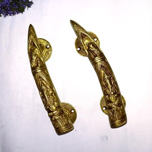 以下が含まれることがあります： 2つの装飾的な金色のドアハンドル。各ハンドルには、人物や模様の精巧なレリーフ彫刻が施されています。ハンドルは湾曲した形状で、ネジ穴のある円形のバックプレートに取り付けられています。全体的なデザインは、ヴィンテージまたはアンティークスタイルを示唆しています。