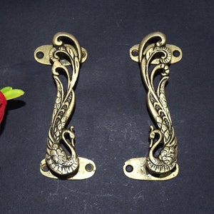 Könnte beinhalten: Zwei verzierte Türklinken aus Messing in Form von Pfauen mit filigranen Details. Die Griffe sind goldfarben und haben ein traditionelles Design.