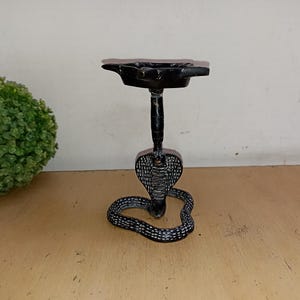 以下が含まれることがあります： 頭が持ち上がったコブラの形をした黒い金属製の灰皿。上部に丸い皿が付いている。