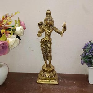 May include: A golden-colored statue of a standing figure, likely a deity, with intricate details. The figure holds objects in both hands and is adorned with ornate clothing and a headdress. The statue is mounted on a decorative base.