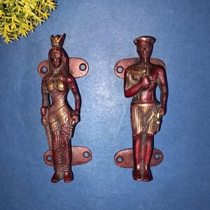 Puede incluir: Un par de tiradores de puerta decorativos con un acabado rojo y dorado. Los tiradores tienen forma de figuras de una mujer y un hombre, ambos vestidos con ropa elaborada.