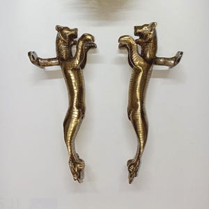 Pode incluir: Duas maçanetas de porta de cor bronze, cada uma em forma de leão em pé com os braços estendidos. As maçanetas têm texturas detalhadas e são montadas em uma superfície branca. A cabeça do leão está voltada para a frente.