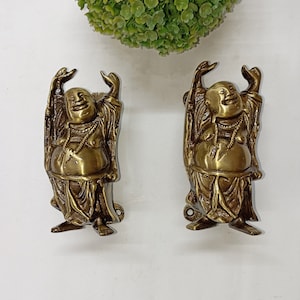 May include: Two gold-toned metal door handles shaped like the laughing Buddha with raised arms. The handles are detailed with intricate carvings and have a traditional Asian design.