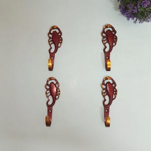 May include: Four decorative wall hooks with a scorpion design. The hooks are a deep red color with gold-colored accents and a curved hook. The hooks are made of metal and are mounted on a light-colored wall.