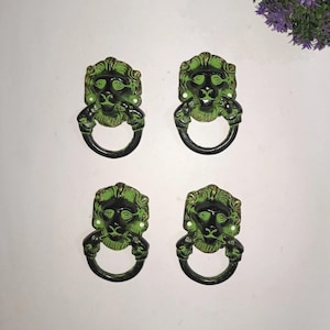 Pode incluir: Um conjunto de quatro puxadores de gaveta de metal, cada um apresentando um design de cabeça de leão com acabamento em preto e verde. Cada puxador inclui um anel circular preso à boca do leão, com orifícios de montagem visíveis.