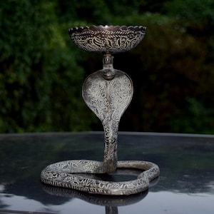 May include: An ornate silver-coloured cobra-shaped candle holder. The candle holder has a detailed design, with a bowl-shaped top and a cobra head and body forming the base. The cobra's body is coiled, and the entire piece has an antique finish.