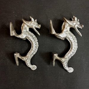 以下が含まれることがあります： 銀色のドラゴンの形をしたキャビネットプルが2つ。各プルは、開いた口と湾曲した体を持つ詳細なドラゴンデザインが特徴です。プルは黒い表面に取り付けられています。